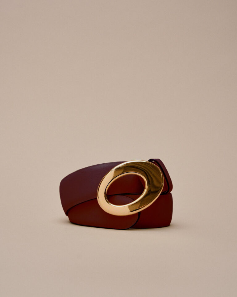 Cinturón estructurado rojo con hebilla dorada sobre fondo beige. © RFB