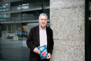 Juan Infante sosteniendo un libro en Bilbao frente a un edificio moderno.